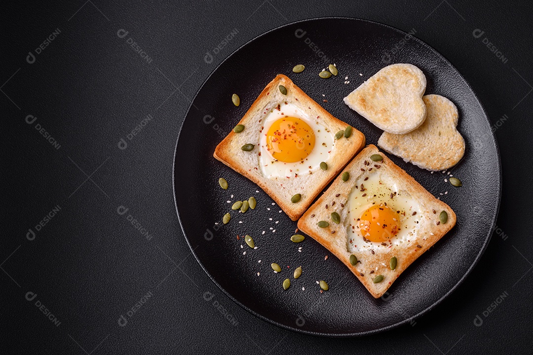 Ovo frito em forma de coração em torradas de pão com sementes de gergelim, sementes de linho e sementes de abóbora em uma placa preta sobre um fundo escuro de concreto