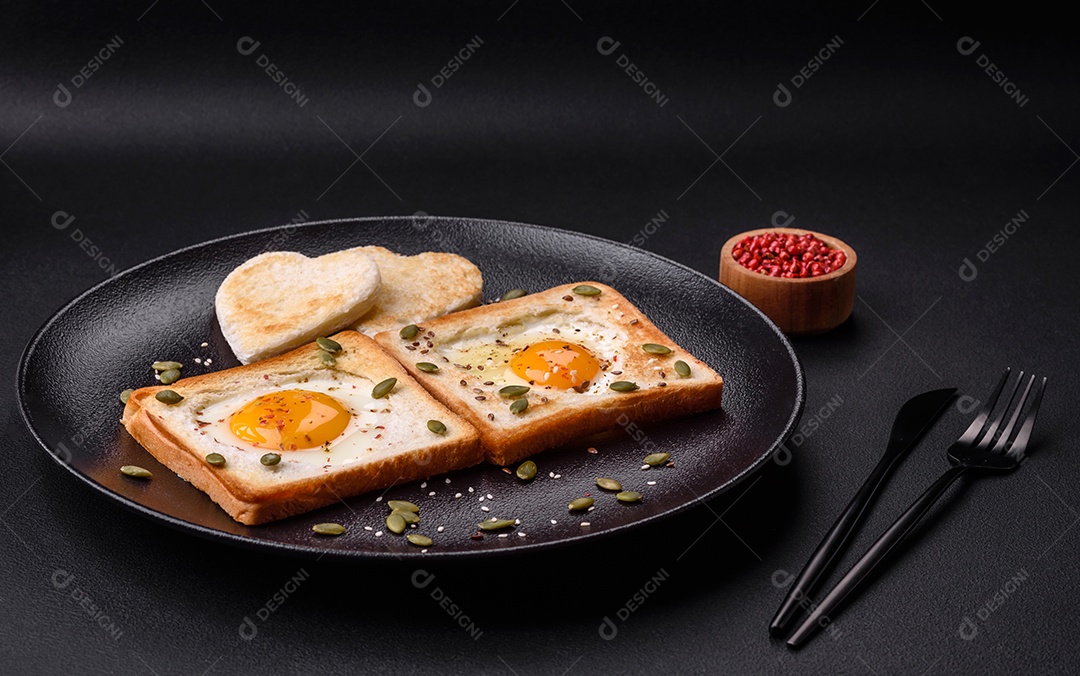 Ovo frito em forma de coração em torradas de pão com sementes de gergelim, sementes de linho e sementes de abóbora em uma placa preta sobre um fundo escuro de concreto