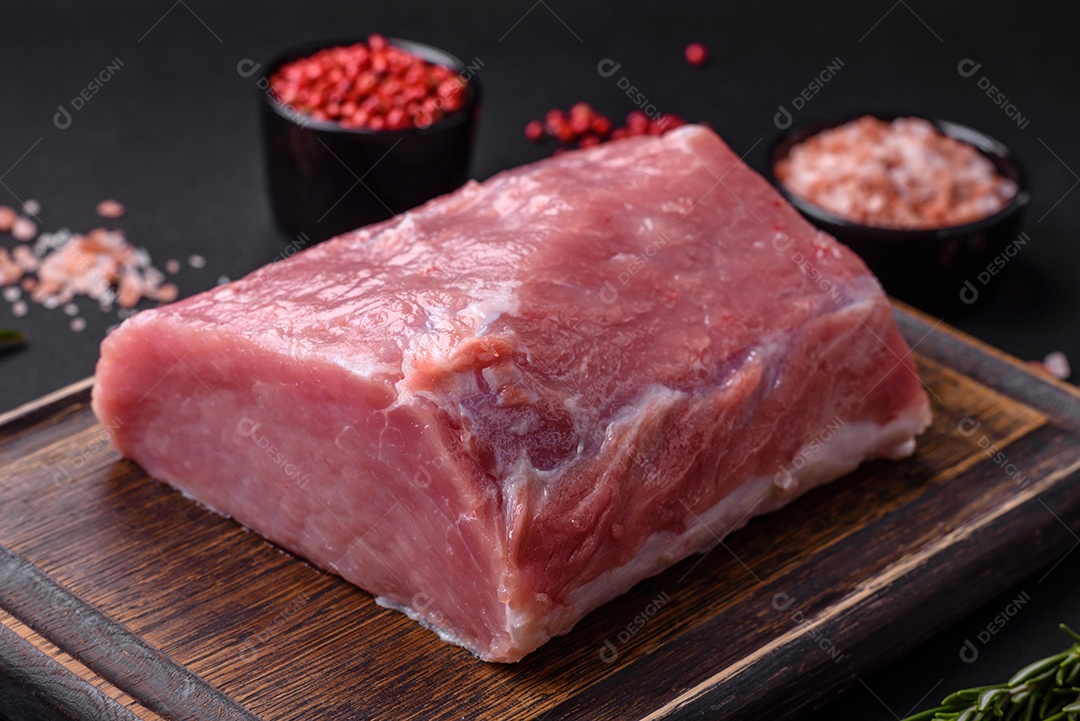Um pedaço de carne de porco fresca crua sobre uma tábua de madeira com especiarias e ervas sobre um fundo escuro de concreto