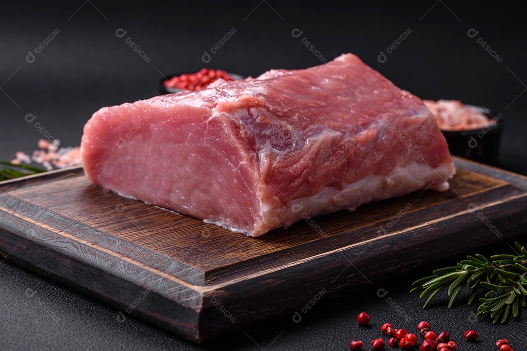Um pedaço de carne de porco fresca crua sobre uma tábua de madeira com especiarias e ervas sobre um fundo escuro de concreto