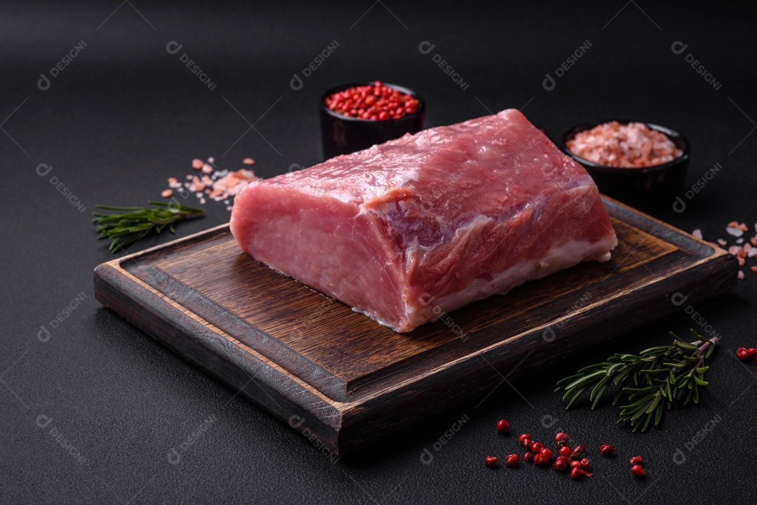 Um pedaço de carne de porco fresca crua sobre uma tábua de madeira com especiarias e ervas sobre um fundo escuro de concreto