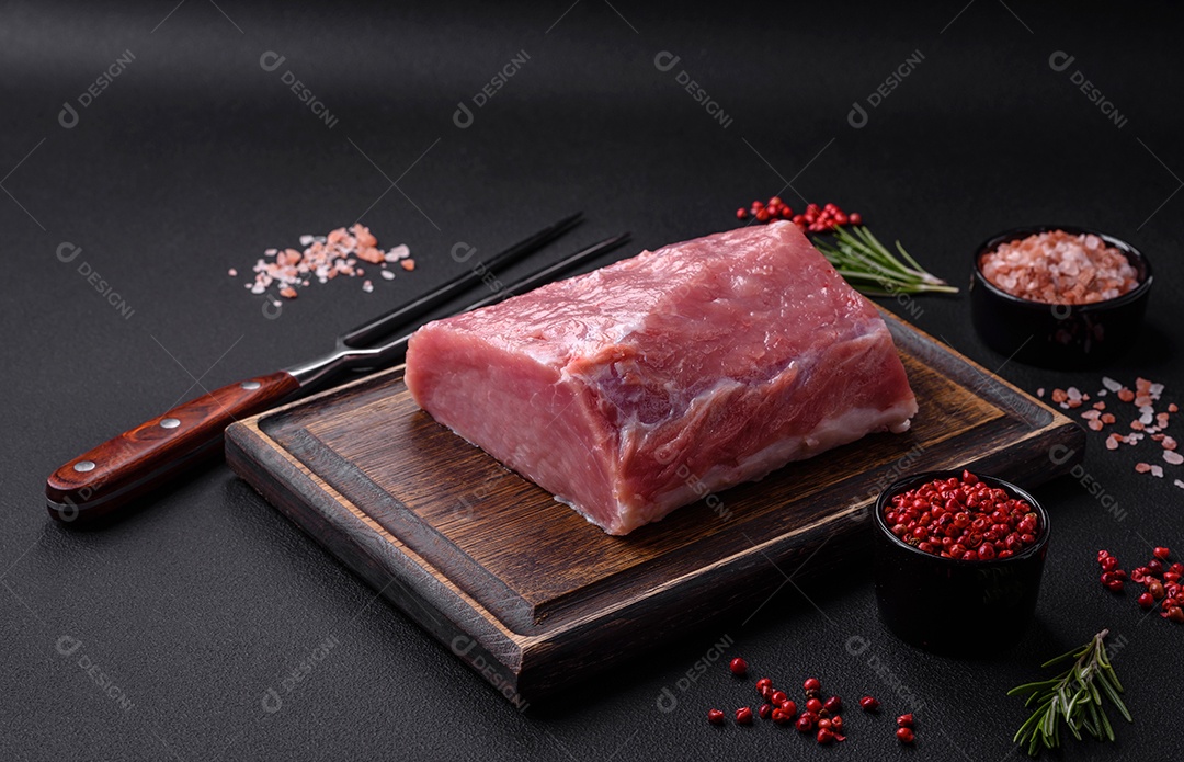 Um pedaço de carne de porco fresca crua sobre uma tábua de madeira com especiarias e ervas sobre um fundo escuro de concreto