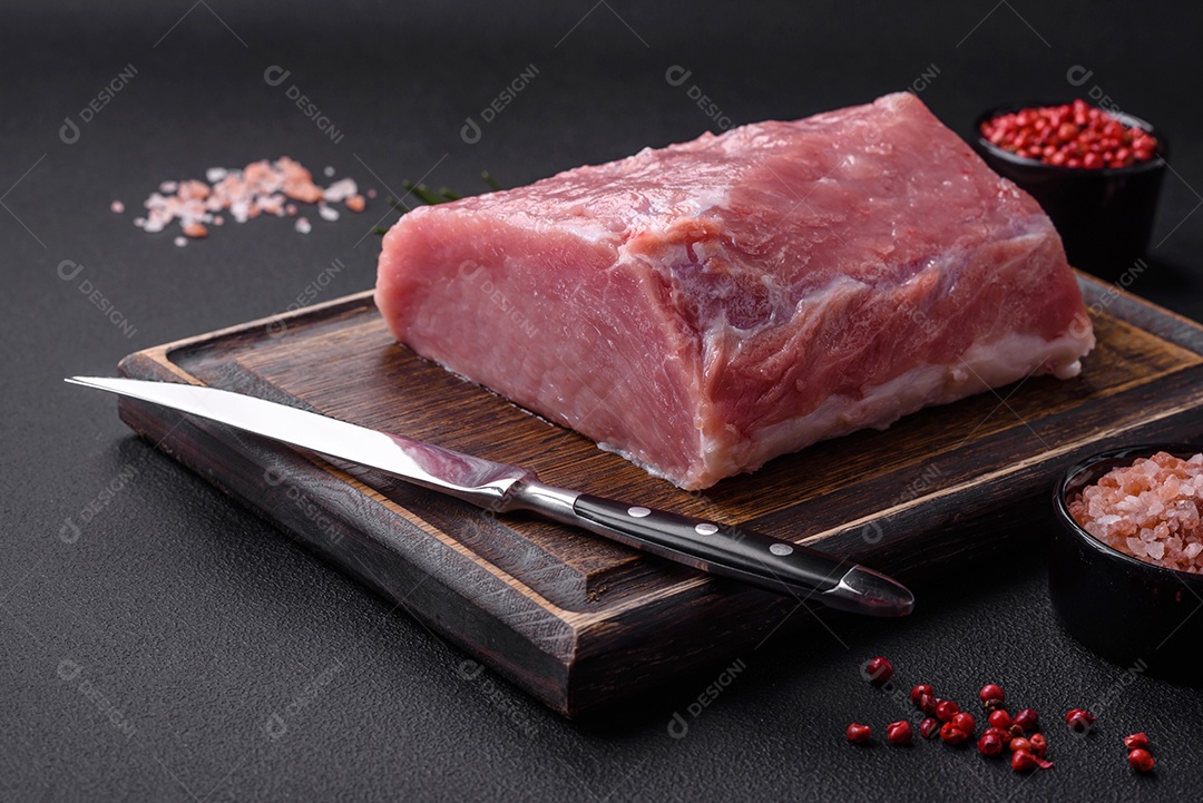 Um pedaço de carne de porco fresca crua sobre uma tábua de madeira com especiarias e ervas sobre um fundo escuro de concreto