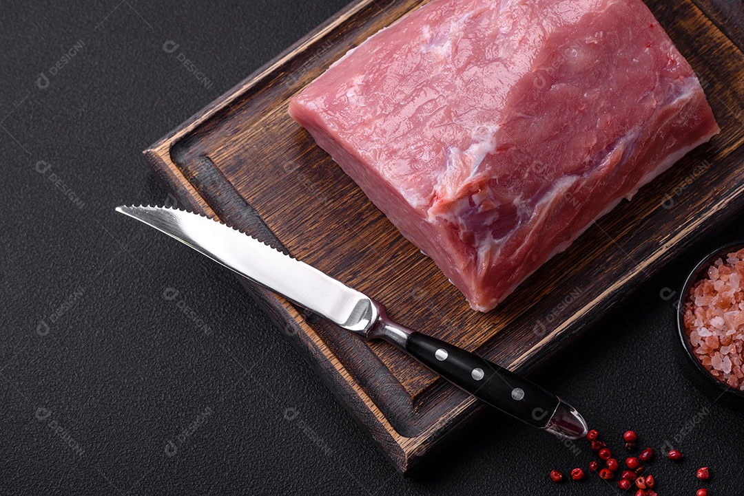 Um pedaço de carne de porco fresca crua sobre uma tábua de madeira com especiarias e ervas sobre um fundo escuro de concreto