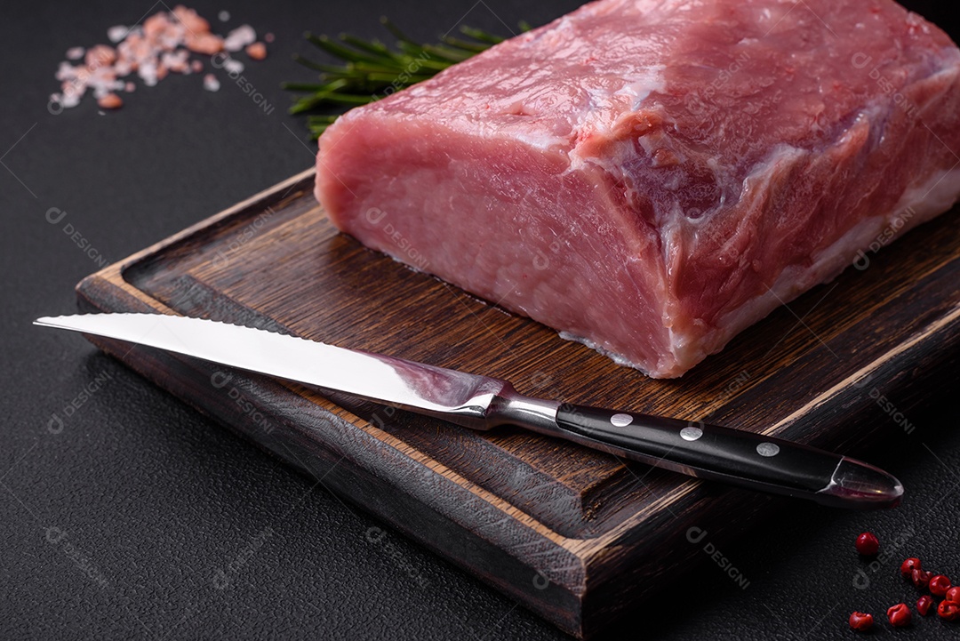 Um pedaço de carne de porco fresca crua sobre uma tábua de madeira com especiarias e ervas sobre um fundo escuro de concreto