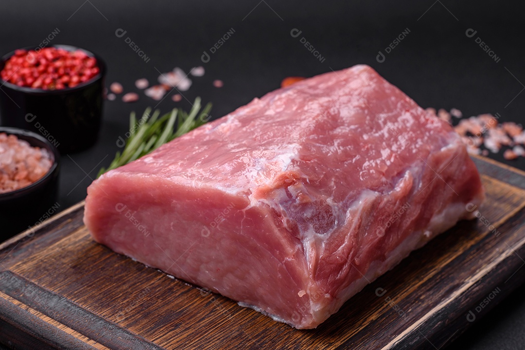 Um pedaço de carne de porco fresca crua sobre uma tábua de madeira com especiarias e ervas sobre um fundo escuro de concreto