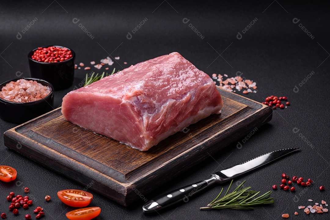 Um pedaço de carne de porco fresca crua sobre uma tábua de madeira com especiarias e ervas sobre um fundo escuro de concreto