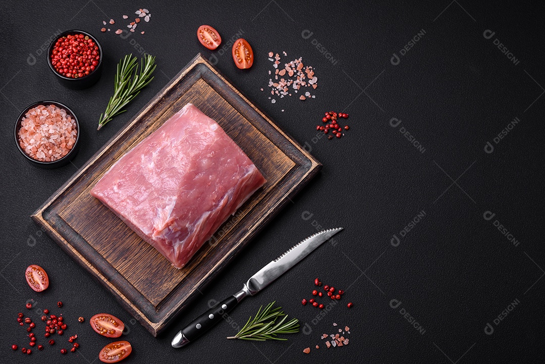 Um pedaço de carne de porco fresca crua sobre uma tábua de madeira com especiarias e ervas sobre um fundo escuro de concreto