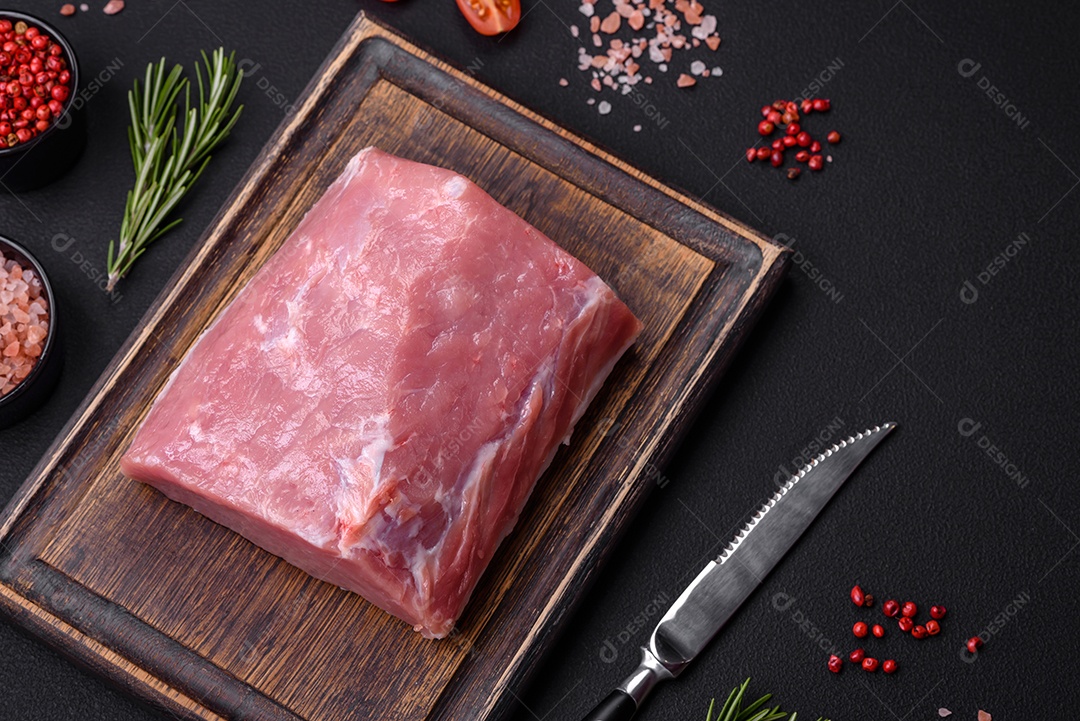 Um pedaço de carne de porco fresca crua sobre uma tábua de madeira com especiarias e ervas sobre um fundo escuro de concreto