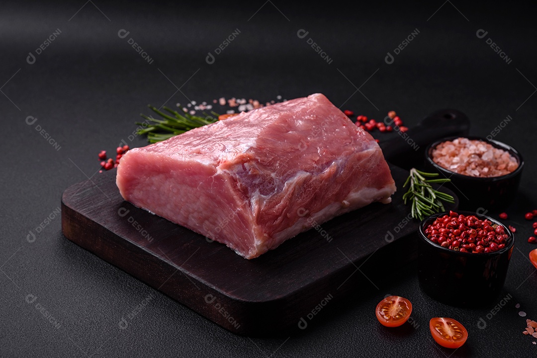 Um pedaço de carne de porco fresca crua sobre uma tábua de madeira com especiarias e ervas sobre um fundo escuro de concreto