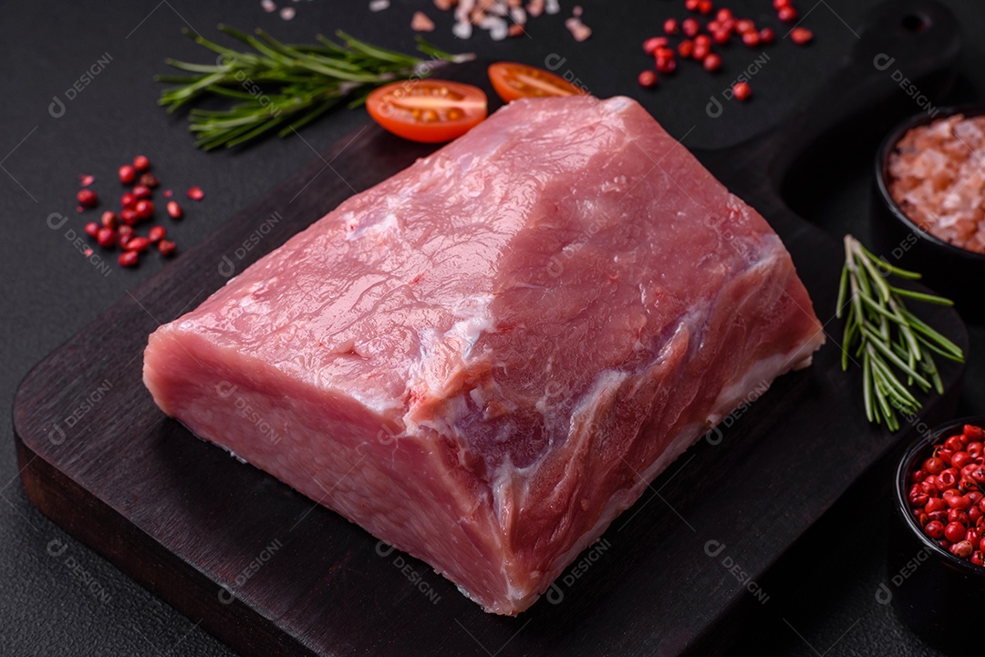 Um pedaço de carne de porco fresca crua sobre uma tábua de madeira com especiarias e ervas sobre um fundo escuro de concreto