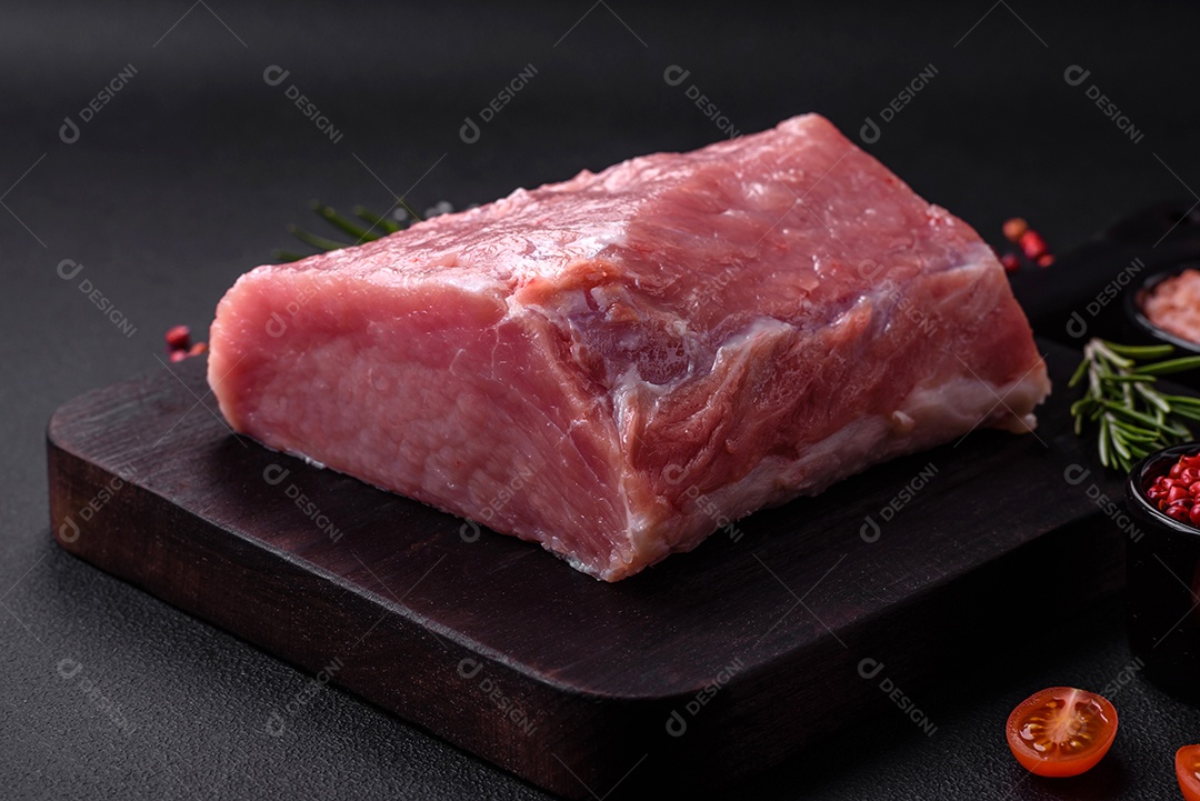 Um pedaço de carne de porco fresca crua sobre uma tábua de madeira com especiarias e ervas sobre um fundo escuro de concreto