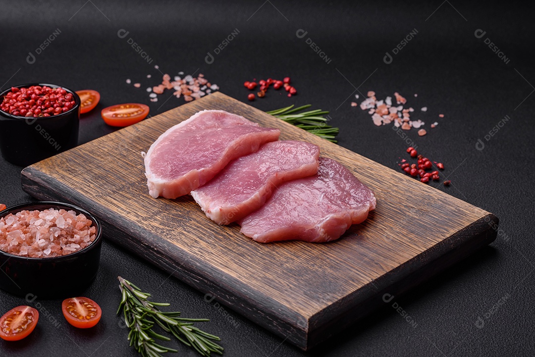 Um pedaço de carne de porco fresca crua sobre uma tábua de madeira com especiarias e ervas sobre um fundo escuro de concreto