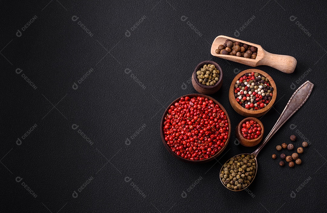 Composição, conceito, consistindo em vários tipos de cores diferentes de pimenta da Jamaica em tigelas e colheres sobre fundo escuro de concreto