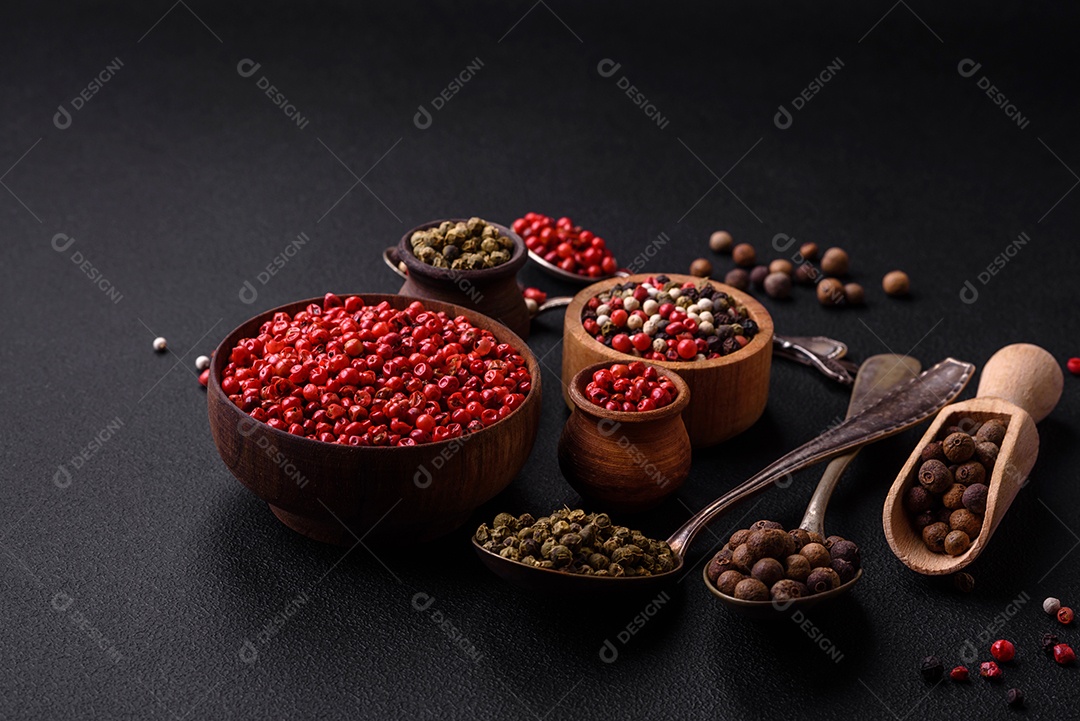 Composição, conceito, consistindo em vários tipos de cores diferentes de pimenta da Jamaica em tigelas e colheres sobre fundo escuro de concreto