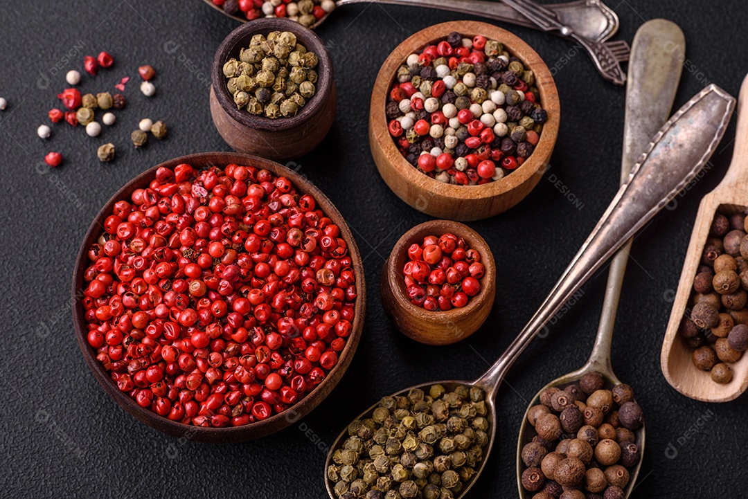 Composição, conceito, consistindo em vários tipos de cores diferentes de pimenta da Jamaica em tigelas e colheres sobre fundo escuro de concreto