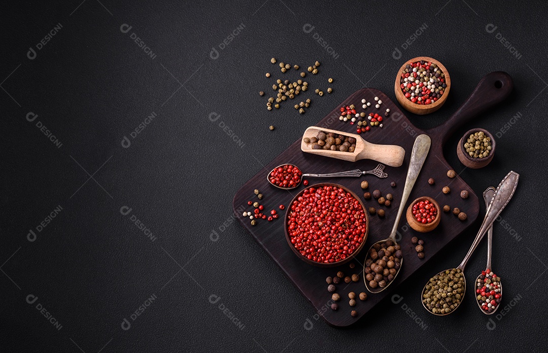 Composição, conceito, consistindo em vários tipos de cores diferentes de pimenta da Jamaica em tigelas e colheres sobre fundo escuro de concreto