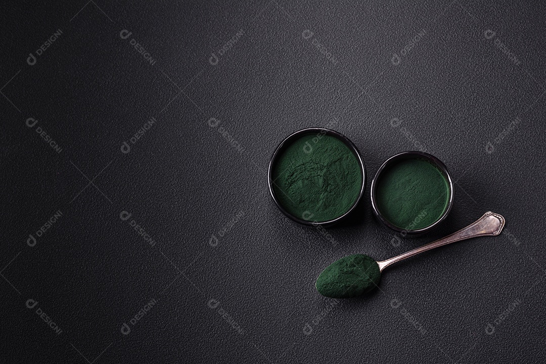 Especiaria, espirulina de cor verde em forma de pó em uma tigela preta sobre um fundo escuro de concreto