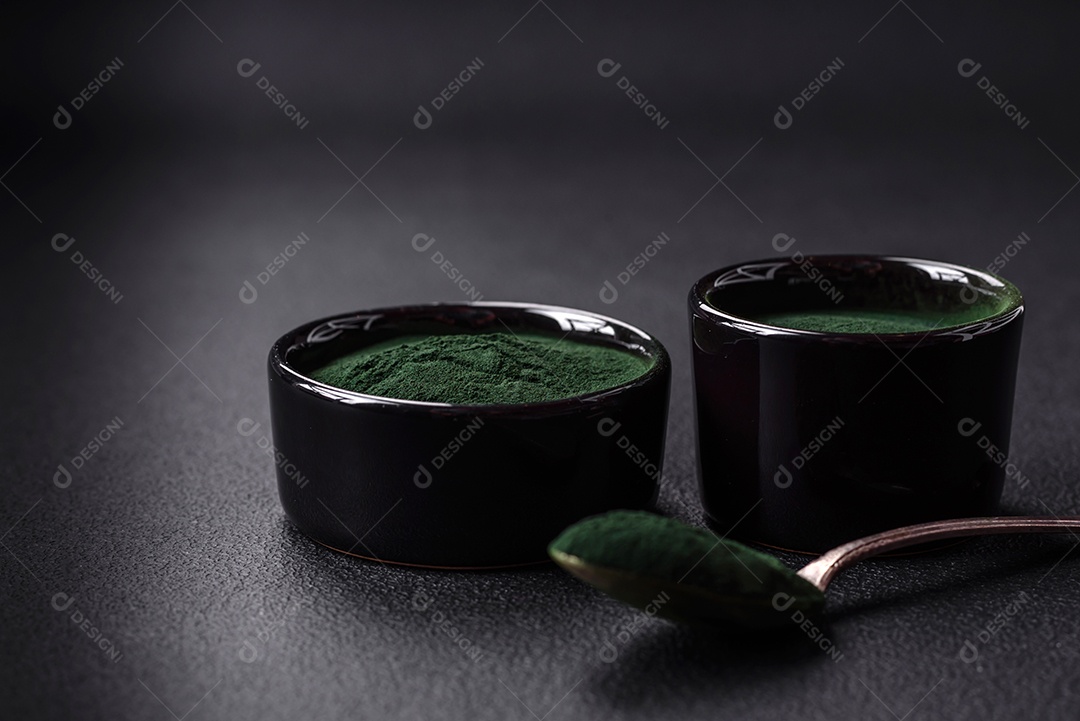 Especiaria, espirulina de cor verde em forma de pó em uma tigela preta sobre um fundo escuro de concreto