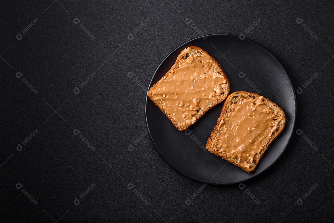 Sanduíche nutritivo composto por pão e manteiga de amendoim em uma placa de cerâmica preta sobre um fundo escuro de concreto