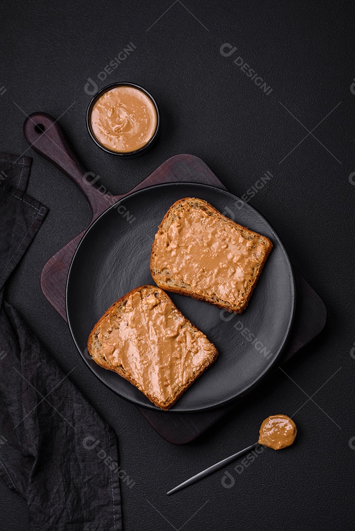 Sanduíche nutritivo composto por pão e manteiga de amendoim em uma placa de cerâmica preta sobre um fundo escuro de concreto