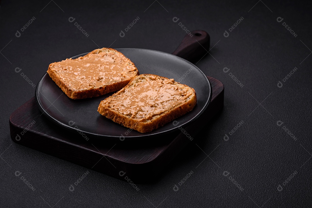 Sanduíche nutritivo composto por pão e manteiga de amendoim em uma placa de cerâmica preta sobre um fundo escuro de concreto