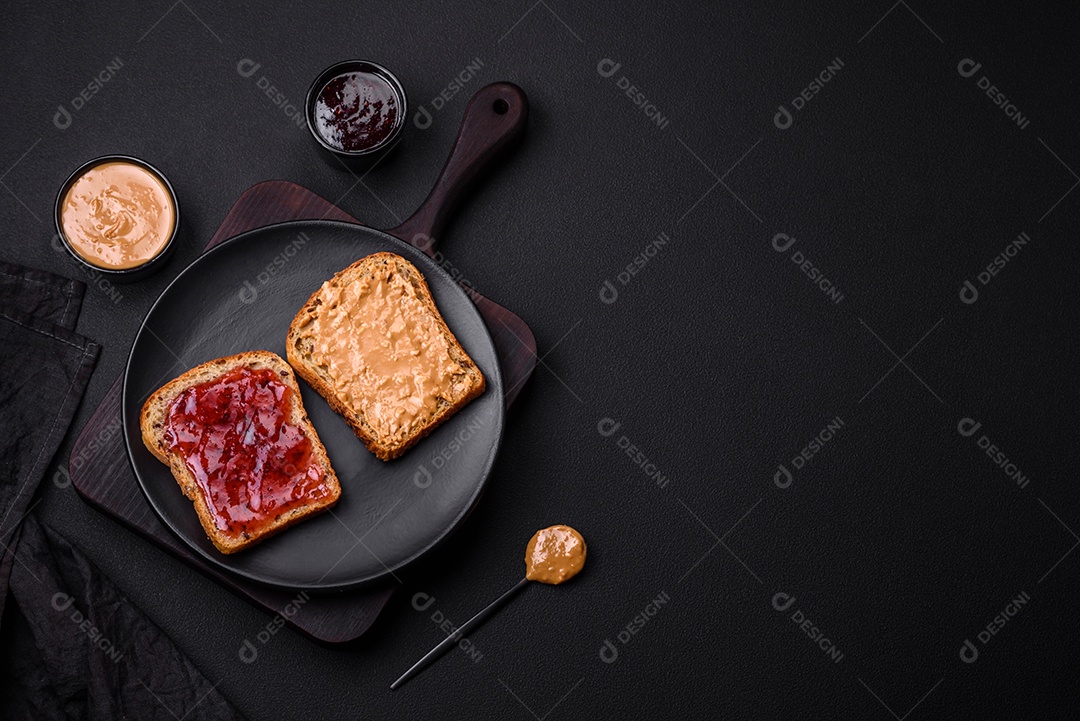 Sanduíches nutritivos compostos por pão, geléia de framboesa e manteiga de amendoim em uma placa de cerâmica preta sobre fundo escuro de concreto