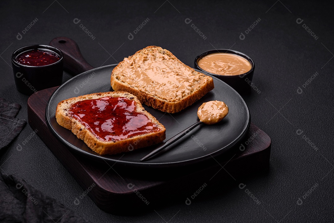 Sanduíches nutritivos compostos por pão, geléia de framboesa e manteiga de amendoim em uma placa de cerâmica preta sobre fundo escuro de concreto