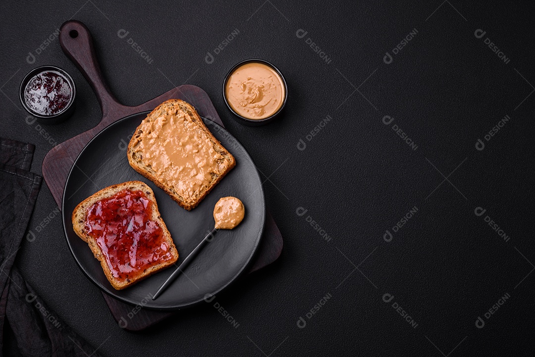 Sanduíches nutritivos compostos por pão, geléia de framboesa e manteiga de amendoim em uma placa de cerâmica preta sobre fundo escuro de concreto