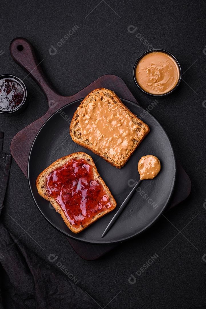 Sanduíches nutritivos compostos por pão, geléia de framboesa e manteiga de amendoim em uma placa de cerâmica preta sobre fundo escuro de concreto