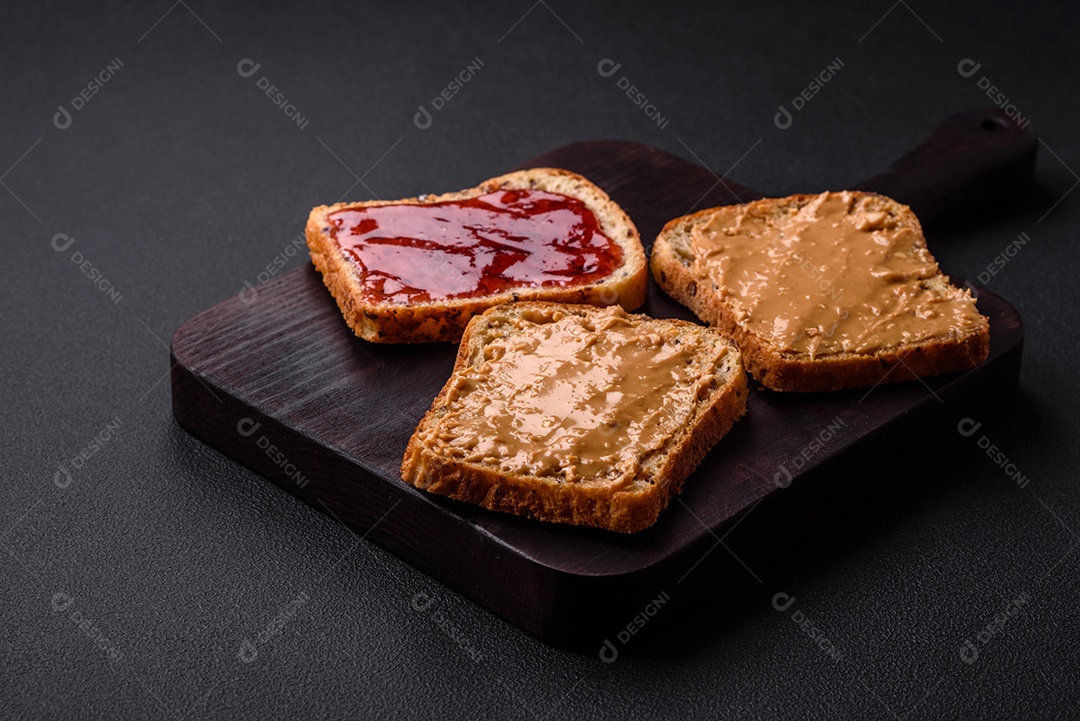 Sanduíches nutritivos compostos por pão, geléia de framboesa e manteiga de amendoim em uma placa de cerâmica preta sobre fundo escuro de concreto