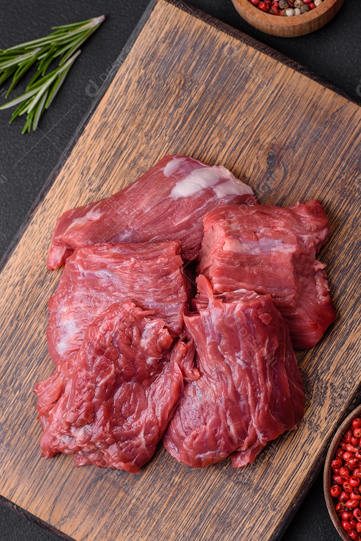 Carne crua cortada em vários pedaços sobre uma tábua de madeira sobre um fundo escuro de concreto