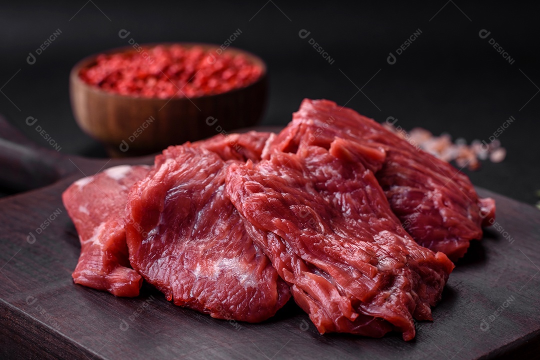 Carne crua cortada em vários pedaços sobre uma tábua de madeira sobre um fundo escuro de concreto