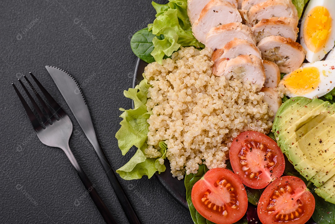 Delicioso almoço saudável composto por frango, abacate, quinoa, ovos, tomate, alface e verduras em uma placa preta sobre fundo de concreto
