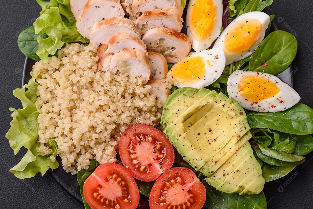 Delicioso almoço saudável composto por frango, abacate, quinoa, ovos, tomate, alface e verduras em uma placa preta sobre fundo de concreto