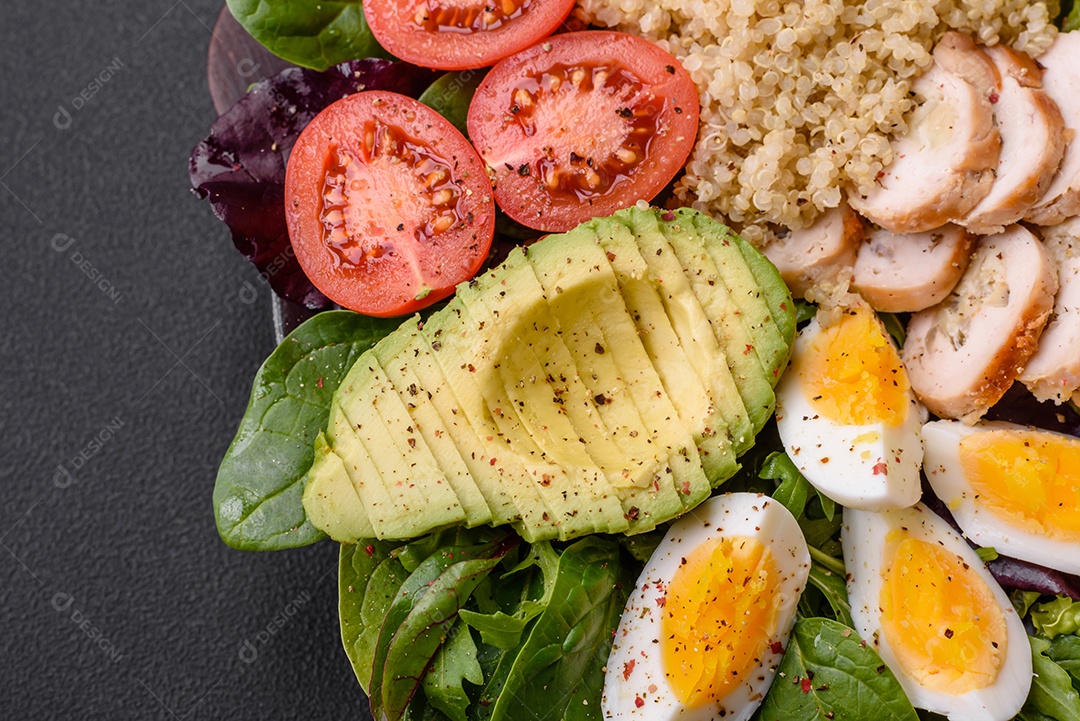 Delicioso almoço saudável composto por frango, abacate, quinoa, ovos, tomate, alface e verduras em uma placa preta sobre fundo de concreto