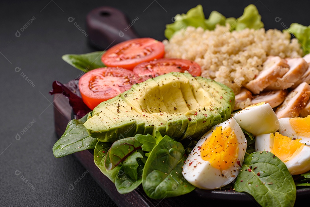 Delicioso almoço saudável composto por frango, abacate, quinoa, ovos, tomate, alface e verduras em uma placa preta sobre fundo de concreto