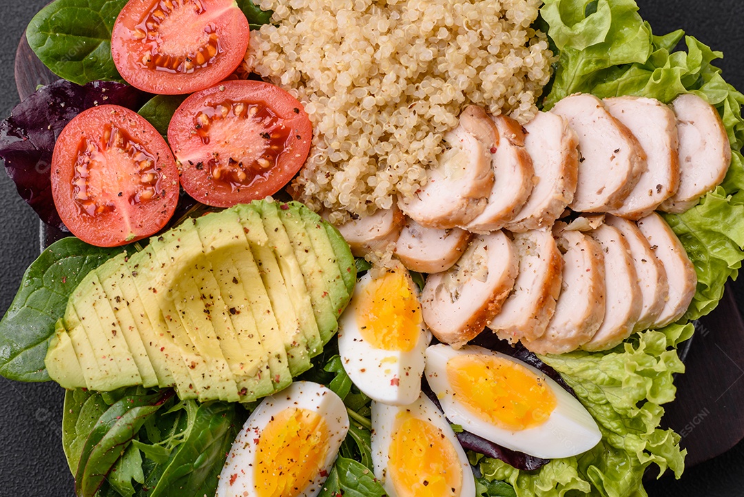 Delicioso almoço saudável composto por frango, abacate, quinoa, ovos, tomate, alface e verduras em uma placa preta sobre fundo de concreto