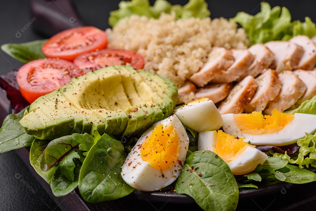 Delicioso almoço saudável composto por frango, abacate, quinoa, ovos, tomate, alface e verduras em uma placa preta sobre fundo de concreto