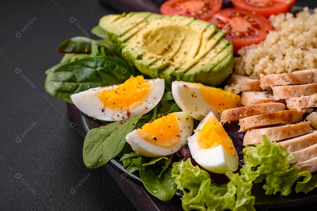 Delicioso almoço saudável composto por frango, abacate, quinoa, ovos, tomate, alface e verduras em uma placa preta sobre fundo de concreto