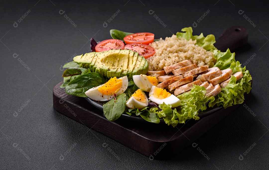 Delicioso almoço saudável composto por frango, abacate, quinoa, ovos, tomate, alface e verduras em uma placa preta sobre fundo de concreto