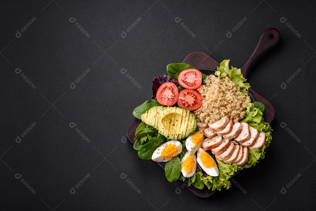Delicioso almoço saudável composto por frango, abacate, quinoa, ovos, tomate, alface e verduras em uma placa preta sobre fundo de concreto