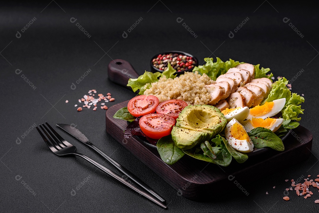 Delicioso almoço saudável composto por frango, abacate, quinoa, ovos, tomate, alface e verduras em uma placa preta sobre fundo de concreto