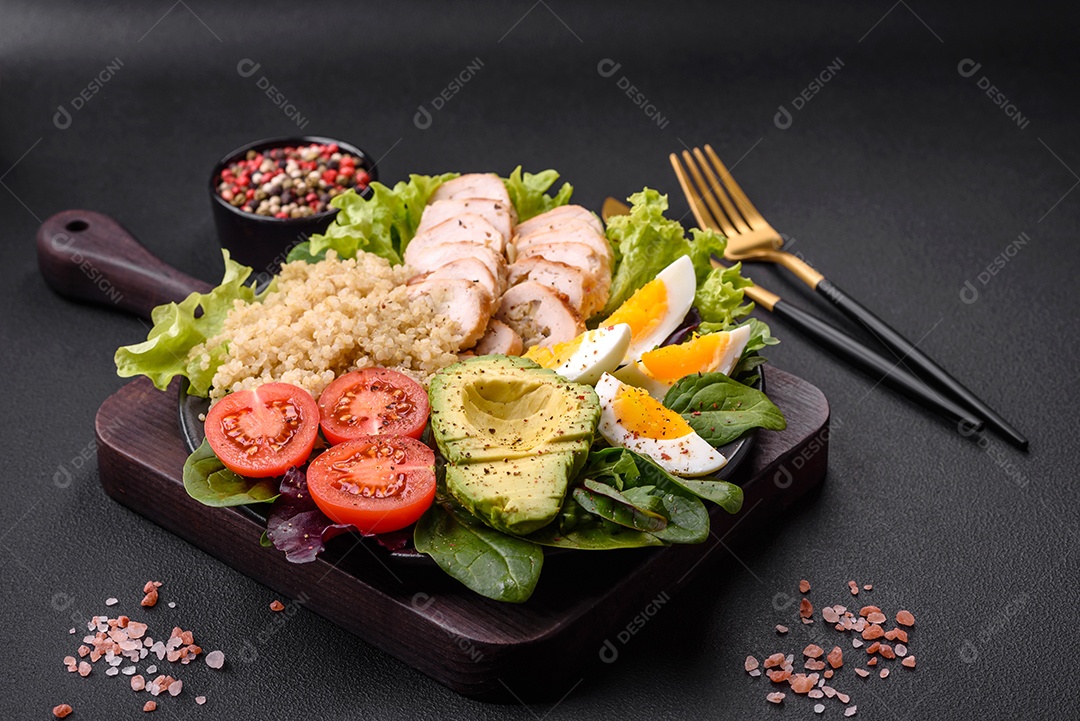 Delicioso almoço saudável composto por frango, abacate, quinoa, ovos, tomate, alface e verduras em uma placa preta sobre fundo de concreto