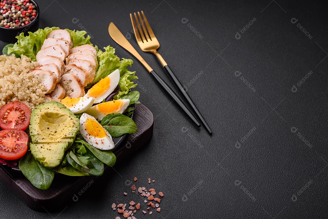 Delicioso almoço saudável composto por frango, abacate, quinoa, ovos, tomate, alface e verduras em uma placa preta sobre fundo de concreto