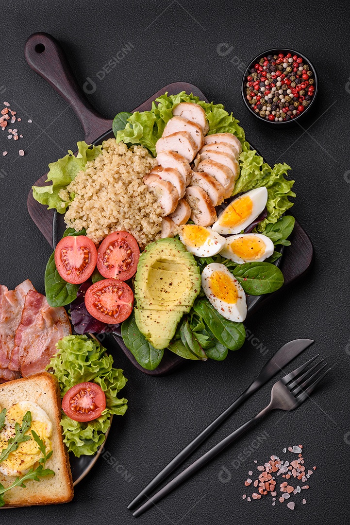 Delicioso almoço saudável composto por frango, abacate, quinoa, ovos, tomate, alface e verduras em uma placa preta sobre fundo de concreto