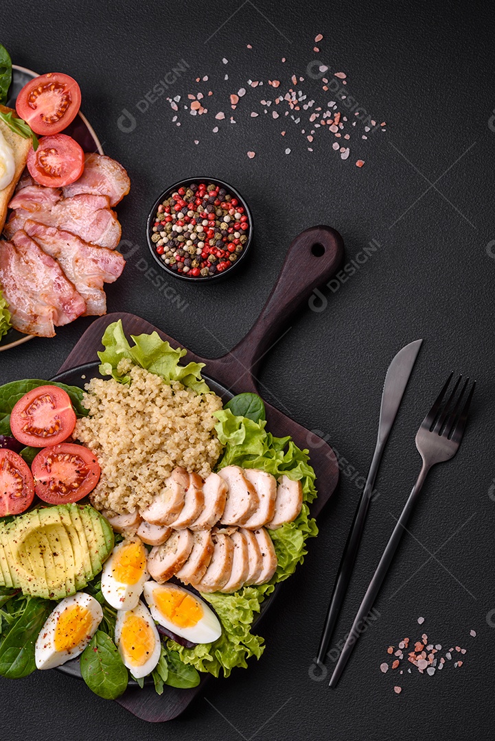 Delicioso almoço saudável composto por frango, abacate, quinoa, ovos, tomate, alface e verduras em uma placa preta sobre fundo de concreto