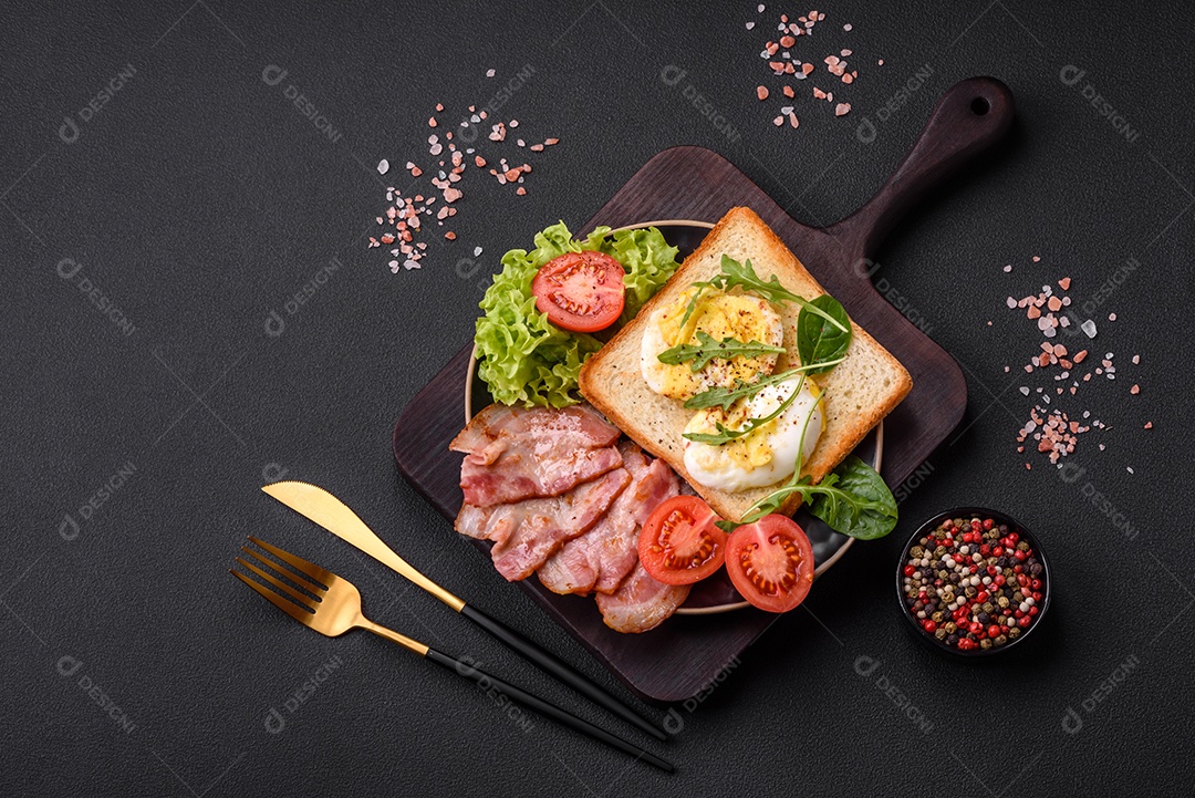 Delicioso almoço saudável composto por bacon, torradas, ovos, tomates, alface e verduras em uma placa preta sobre um fundo de concreto