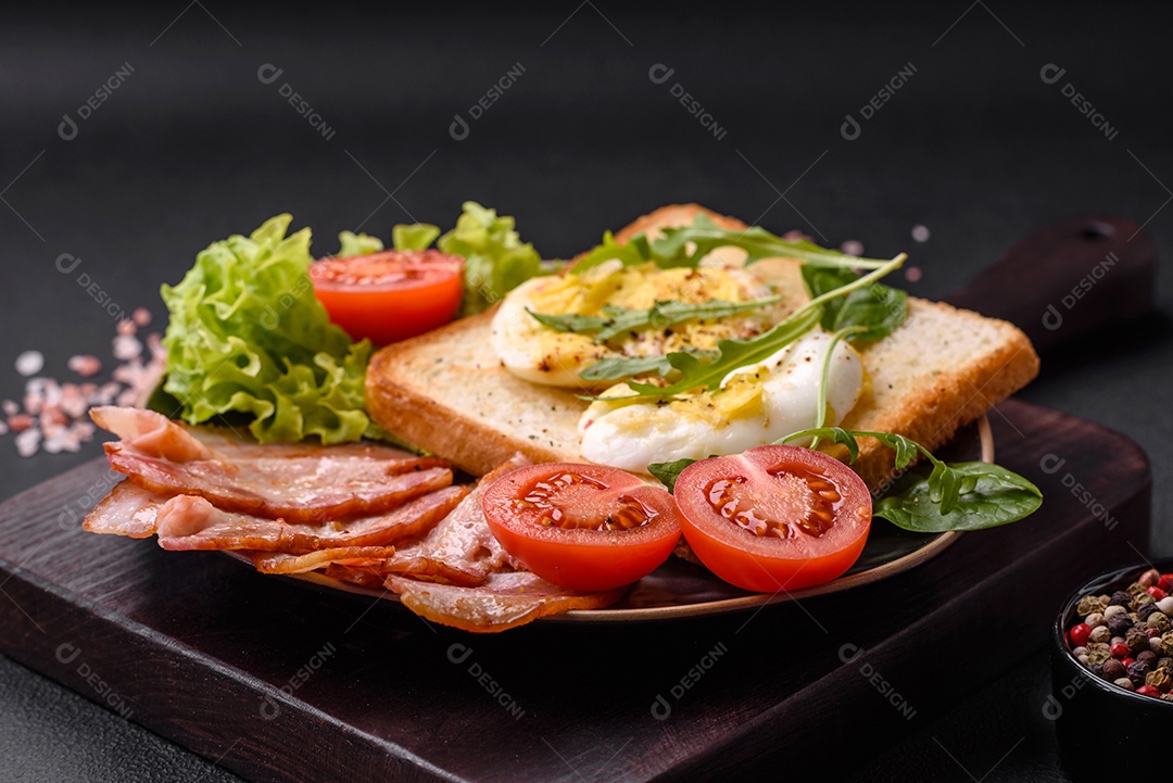 Delicioso almoço saudável composto por bacon, torradas, ovos, tomates, alface e verduras em uma placa preta sobre um fundo de concreto