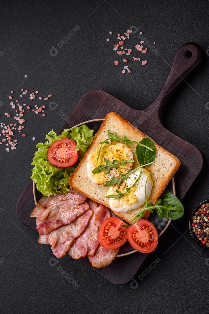 Delicioso almoço saudável composto por bacon, torradas, ovos, tomates, alface e verduras em uma placa preta sobre um fundo de concreto